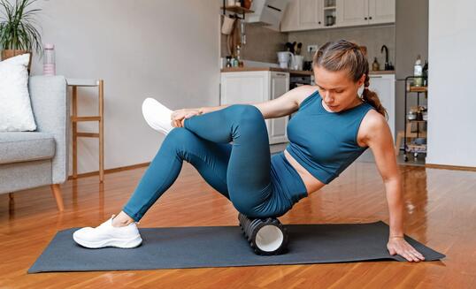 Athletic woman does a workout at home, using a foam roller to release leg muscles. Self-myofascial release.