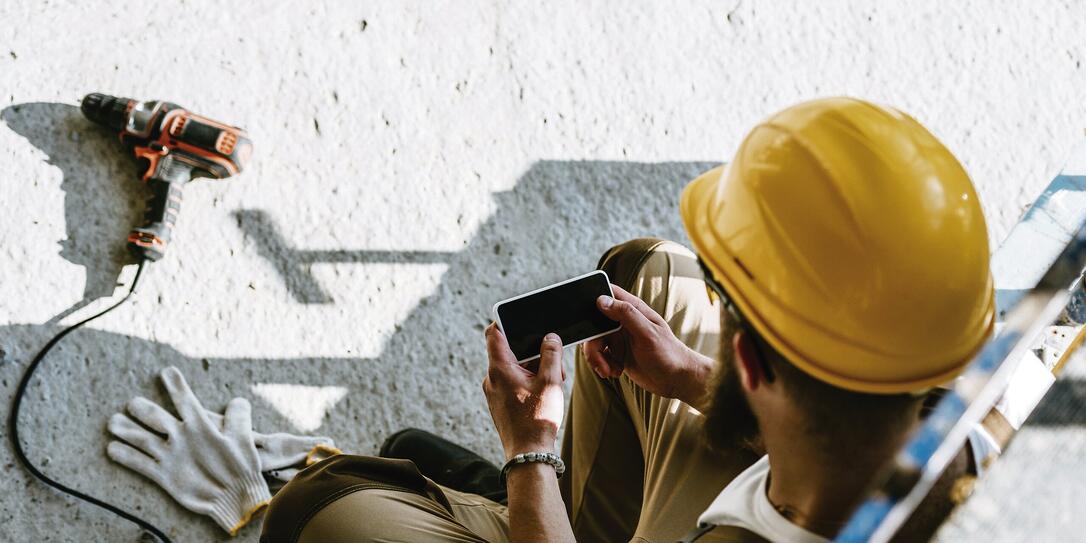 overhead view of builder in protective helmet using smartphone with blank screen at construction site