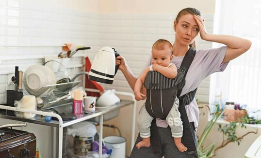 Tired multitasking mother and child in baby carrier. Young girl in the kitchen with a child. The difficulties of motherhood and loneliness