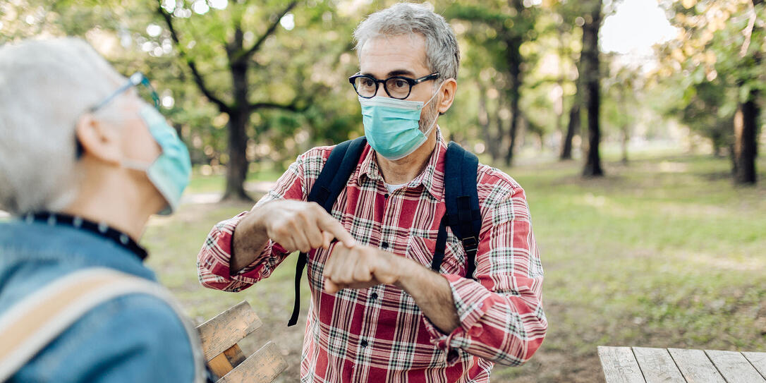 Senior man and his wife with protective face masks resting in the nature and talking on sign language